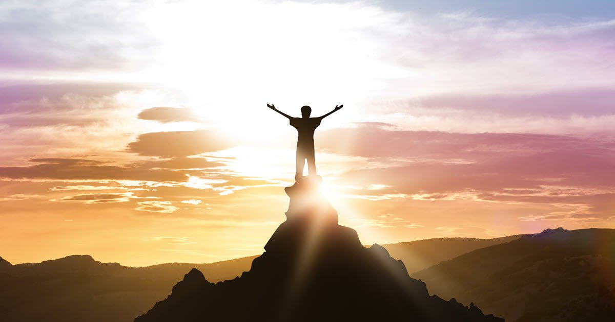 Man on top of the mountains facing the sun in a hero pose.